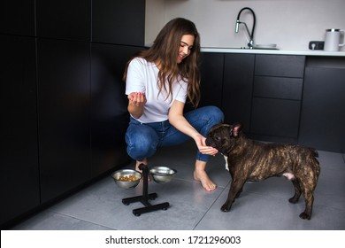 Young Woman In Kitchen During Quarantine. Girl Sit In Squat Pose And Feed French Bulldog. Adult Pet Eating Dog Food. Pet Care.