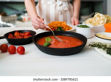 Young woman in kitchen making delicious pasta with sauce using a pan at home. Italian rural cooking still life. Wooden board, fresh vegetables, cooked pasta and a pan of sauce on wooden kitchen table
