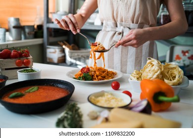 Young Woman In Kitchen Making Delicious Pasta With Sauce Using A Pan At Home. Italian Rural Cooking Still Life. Wooden Board, Fresh Vegetables, Cooked Pasta And A Pan Of Sauce On Wooden Kitchen Table