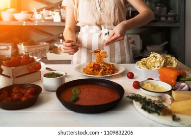 Young Woman In Kitchen Making Delicious Pasta With Sauce Using A Pan At Home. Italian Rural Cooking Still Life. Wooden Board, Fresh Vegetables, Cooked Pasta And A Pan Of Sauce On Wooden Kitchen Table.