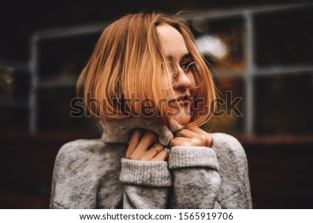 Young woman in a knitted grey turtleneck sweater covering her face. Close up. Blonde girl in glasses basking in grey turtleneck. Outdoor close up portrait of young beautiful woman.