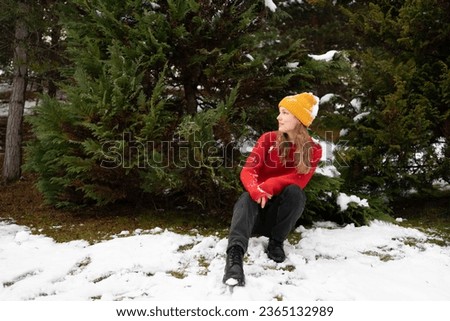 A young woman in a knitted hat rests after playing snowballs against a background of green Christmas trees outdoors. Concept of winter activities