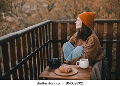 Young woman in knitted sweater and hat drinking tea and eating fresh croissants on cozy balcony of a wooden country house on autumn day. 