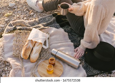 Young woman in knitted sweater having picnic at beach with tea in thermos and fresh baguettes in wicker eco bag. Cozy slow lifestyle concept.