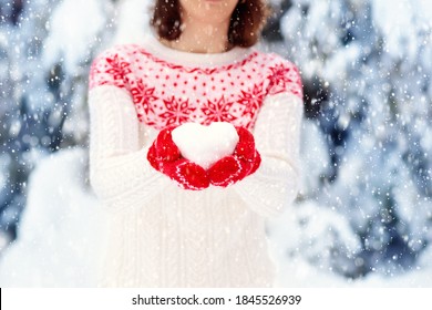 Young Woman In Knitted Sweater Holding Heart Shape Snow Ball In Winter. Girl In Family Snow Fight Game. Female In Knit Handmade Hat And Mittens With Christmas Snowflake Design Having Fun In Snowy Park