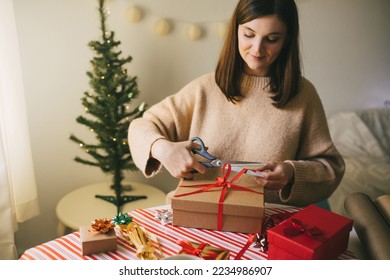 Young woman in knitted sweater wrapping Christmas presents using paper, scissors and colorful ribbons. Festive holiday preparations.