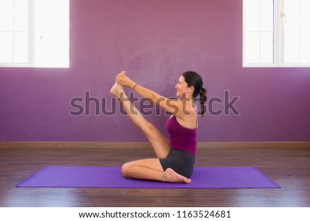 Young woman in krounchasana on purple mat. Female yogi in heron pose on purple studio with big windows. Intense hamstring stretch. Lady with leg up and hands holding foot. Flexibility concept