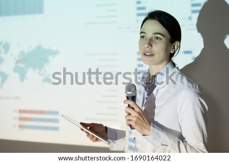 Young woman in lab coat using thesis plan on tablet while presenting project at medical convention