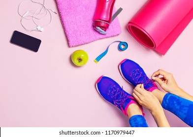 Young woman laces sneakers, preparing for training. Bottle of water, yoga mat, phone, headphones on pastel pink background flat lay top view. Sports shoes, Fitness concept - Powered by Shutterstock