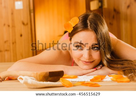 Young woman with ladle and petals relaxing laying in wood finnish sauna. Attractive girl in bikini resting. Spa wellbeing pleasure.