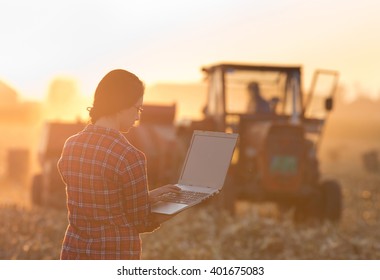 Young woman with laptop standing on field in sunset while tractor baling in background