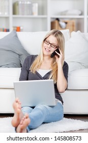 Young Woman With Laptop Using Cordless Phone Against Sofa At Home