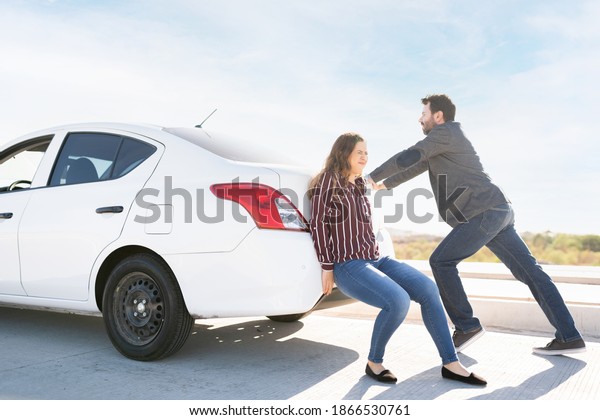 Young woman and latin adult guy pushing their\
broken-down car to the roadside\
