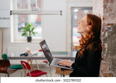 Young woman laughing at a good online joke as she relaxes leaning against an interior brick wall in an office holding her open laptop