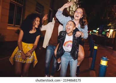 Young Woman Laughing Hysterically While Piggybacking Her Friend. Friends Having A Good Time While Going Out In The City At Night. Group Of Vibrant Friends Hanging Out Together On The Weekend.