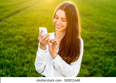 Young woman laughing and typing message in smart phone in the green field during spring or summer 