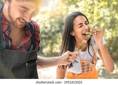 a young woman laughs as she eats skewers of meat from her friend's hands during a trip to the countryside. Lifestyle concept of young adults having fun spending time together with fun activities