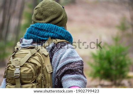 Young woman with layers of heavy, colored winter clothes, blue scarf and green hat from behind, during a walk or a hike in the woodlands.