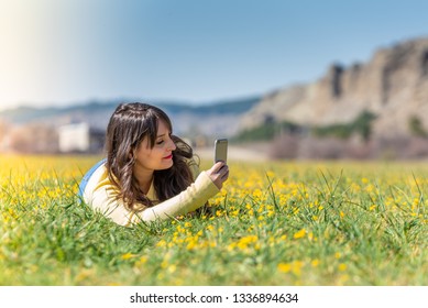 Young Woman Laying Down On A Field Of Yellow Flowers Using Her Mobile Phone. Using Cell Phone Into The Nature. Lifestyle Concept.