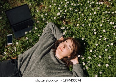 Young Woman Laying In Green Grass Next To The Laptop. People Fatigue From Work. Sleeping And Relaxation Techniques. Vitamin D Sunbathing. Female Power Nap With Eye Closed. Rest After Work From Home