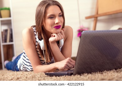 A young woman laying on the floor in front of her laptop with headphones, she is studying or working at home.