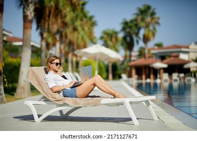 A Young Woman Is Laying On The Sun Lounger Near The Pool With A Laptop. Outdoor