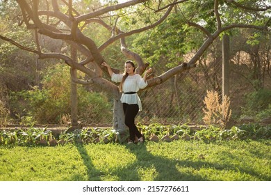 Young Woman Leaning Against A Plum Tree. Full Length Portrait Of A Mexican Woman Leaning Against A Plum Tree At Sunset.