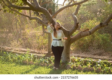 Young Woman Leaning Against A Plum Tree. Full Length Portrait Of A Mexican Woman Leaning Against A Plum Tree At Sunset.