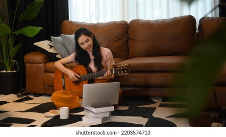 Young Woman Learning To Play Guitar, Using Laptop Computer On Carpet In Living Room