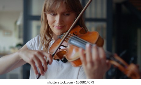 Young woman learning to play violin at home. Romantic girl playing violin with bow. Portrait of female musician performing on string instrument. Dreamy violinist fingers pressing strings on violin