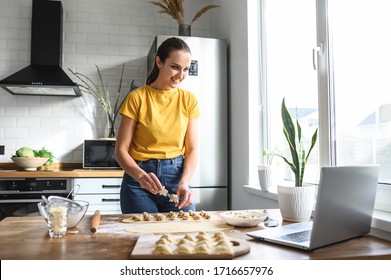 A Young Woman Learns To Cook, She Watches Video Recipes On A Laptop In The Kitchen And Cook A Dish From The Dough. Cooking At Home Concept