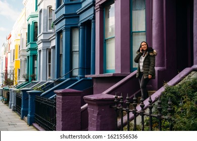 Young Woman Leaving Her House And Using Her Smart Mobile With Colorful Houses On Portobello Street In The Notting Hill Neighborhood