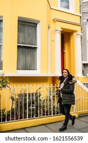 Young Woman Leaving Her House And Using Her Smart Mobile With Colorful Houses On Portobello Street In The Notting Hill Neighborhood