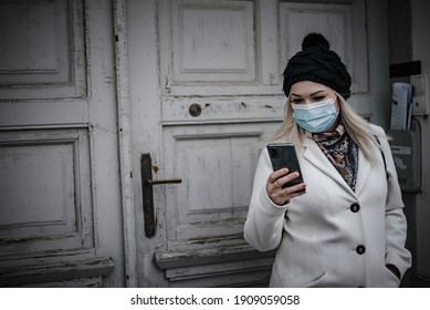 Young Woman Leaving House In Winter, Reading Messages And Wearing Face Mask Due To Covid-19 Pandemic