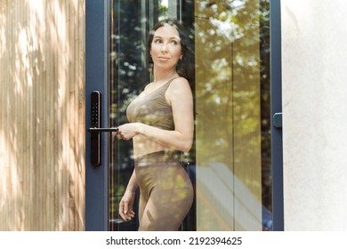 Young Woman Leaving The Modern Country House, Looking Through The Glass Door, Enjoying Beautiful Outdoors Atmosphere.