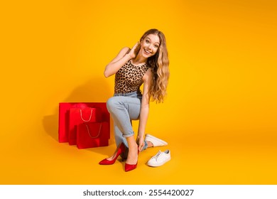 Young woman in leopard print top smiles putting on red high heels against vibrant yellow background, surrounded by shopping bags - Powered by Shutterstock