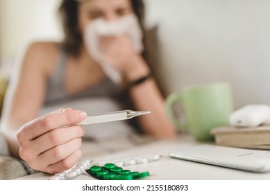 A young woman lies in bed and blows her nose into a handkerchief, takes her temperature and reaches for the pills with her hand. Self-isolation and sick leave at home. Cold
