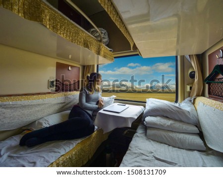 Young woman lies on the bed and looks through the window of the sleeper train crossing picturesque Tibet. Female tourist observing the landscape while travelling along the Trans-Himalayan railway.