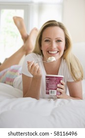 A Young Woman Lies On Her Bed While Eating A Pint Of Ice Cream. She Is Smiling At The Camera.  Vertical Shot.