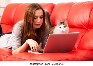 Young Woman Lies On The Red Sofa With Cat And Working Laptop