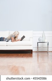 Young Woman Lies On A White Sofa In A Checkered Top And Blue Jeans While Reading A Book. Vertical Shot.