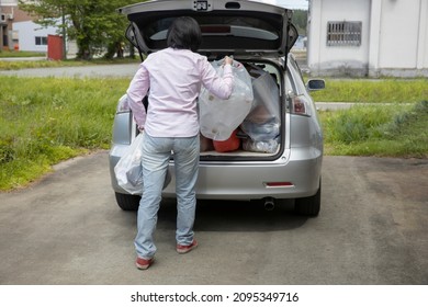 A Young Woman Is Lifting Trash Bag And  Into Trunk Of A Car