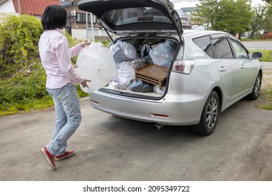 A Young Woman Is Lifting Trash Bag And  Into Trunk Of A Car