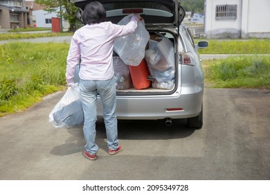 A Young Woman Is Lifting Trash Bag And  Into Trunk Of A Car