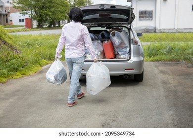 A Young Woman Is Lifting Trash Bag And  Into Trunk Of A Car