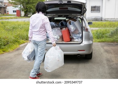 A Young Woman Is Lifting Trash Bag And  Into Trunk Of A Car