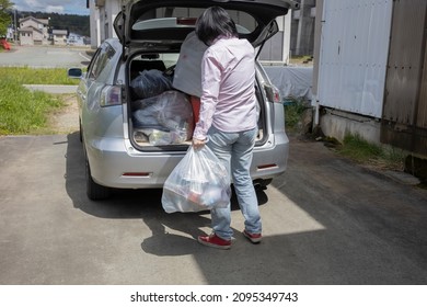 A Young Woman Is Lifting Trash Bag And  Into Trunk Of A Car