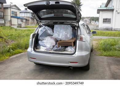 A Young Woman Is Lifting Trash Bag And  Into Trunk Of A Car