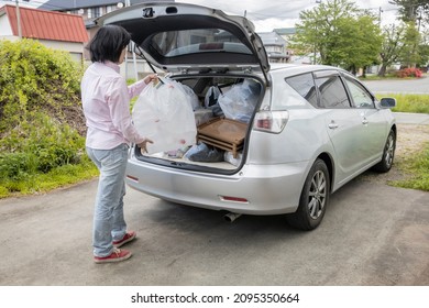 A Young Woman Is Lifting Trash Bag And  Into Trunk Of A Car