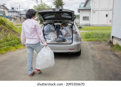 A Young Woman Is Lifting Trash Bag And  Into Trunk Of A Car